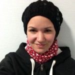 Karina sitzt im Wartezimmer eines Arztes. Sie trägt eine Beanie Mütze, eine Halskrause mit einem Überzug der Sternchen auf rotem Hintergrund zeigt, und einem Sweater der "EDS" als Schriftzug zeigt.