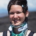 Karina, a woman with short brown hair is smiling, her eyes small from the sun in her face. She is wearing a neck brace and her flower tattoo on her shoulder is visible