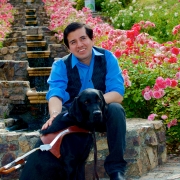 Belo Cipriani, ein Mann der ein blaues Shirt trägt sitzt auf einer steinernen Treppe in Mitten eines Blumenmeers mit pinken Blumen. Vor ihm sitzt sein Assistenzhund, ein Labrador, den er mit der rechten Hand hält.