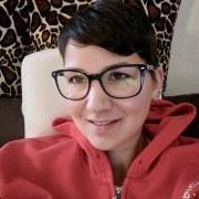 Eine Frau mit kurzen, braunen Haaren und großer, schwarzer, eckiger Brille sitzt auf einem braunen Sofa und trägt einen roten Sweater. Im Hintergrund sind Kissen mit Giraffenmusterung