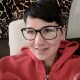Eine Frau mit kurzen, braunen Haaren und großer, schwarzer, eckiger Brille sitzt auf einem braunen Sofa und trägt einen roten Sweater. Im Hintergrund sind Kissen mit Giraffenmusterung