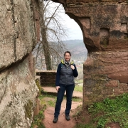 Eine junge Frau steht in einem Durchgang einer felsernen Wand. Sie trägt Wanderbekleidung, einen Rucksack und lächelt. Hinter ihr sieht man blätterlose Bäume.