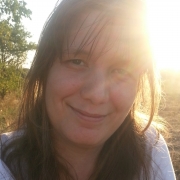Close-up von Cindy, die braune, lange Haare hat, die locker um ihr Gesicht fallen. Sie ist in der Natur und lächelt. Im Hintergrund scheint die Sonne.