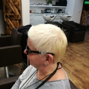 Andrea Eberl, eine Frau mit kurzen, hell-blonden Haaren sitzt auf einem Friseurstuhl und trägt eine schwarze Sonnenbrille - Seitenaufnahme