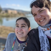 Caitlin, eine junge Frau mit langen braunen Haaren, die gerne alles in Regenbogenfarben trägt, steht neben Karina, einer Frau mit kurzen, braunen Haaren, die einen bunten Schal trägt. Im Hintergrund ist ein breiter Fluss. Die Sonne scheint.
