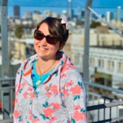 Karina steht auf dem Dach eines Hauses mit Downtown San Francisco im Hintergrund. Sie hat kurze braune Haare und trägt eine rote Blume im Haar.
