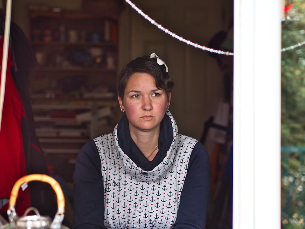 Karina, eine Frau mit kurzen, braunen Haaren und einer roten Blume im Haar sitzt an einem Tisch und schaut aus dem Fenster. Sie trägt einen blauen Pullover mit Kragen.