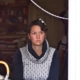 Karina, eine Frau mit kurzen, braunen Haaren und einer roten Blume im Haar sitzt an einem Tisch und schaut aus dem Fenster. Sie trägt einen blauen Pullover mit Kragen.