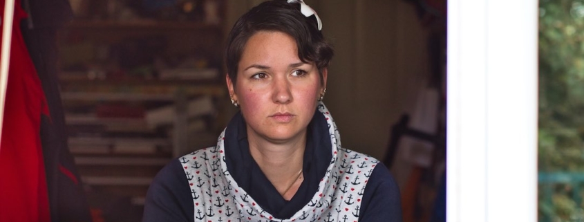 Karina, eine Frau mit kurzen, braunen Haaren und einer roten Blume im Haar sitzt an einem Tisch und schaut aus dem Fenster. Sie trägt einen blauen Pullover mit Kragen.