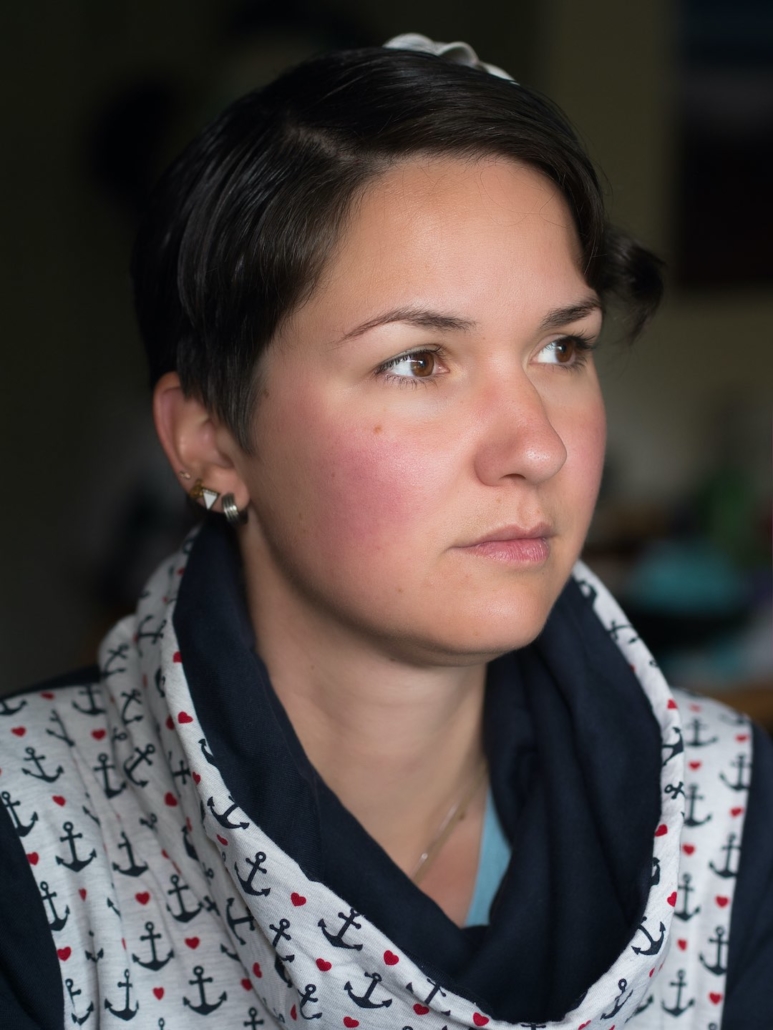 Karina, eine Frau mit kurzen, braunen Haaren und einer roten Blume im Haar sitzt an einem Tisch und schaut aus dem Fenster. Sie trägt einen blauen Pullover mit Kragen.
