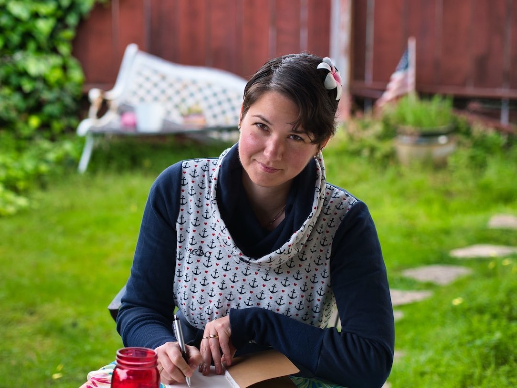 Karina, eine Frau mit kurzen, braunen Haaren und einer roten Blume im Haar sitzt in einem Garten und schreibt in ein Notizbuch.