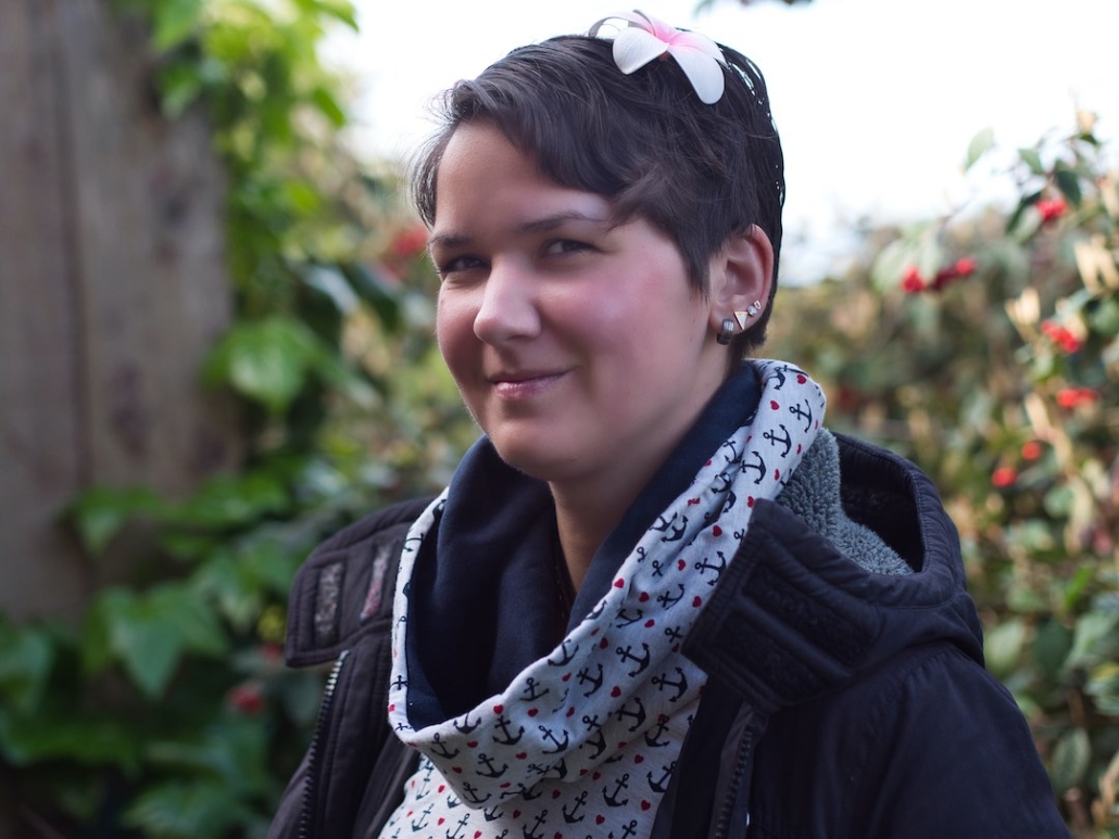 Karina, eine Frau mit kurzen, braunen Haaren und einer roten Blume im Haar steht in einem Garten vor grünem Efeu und lächelt leicht.