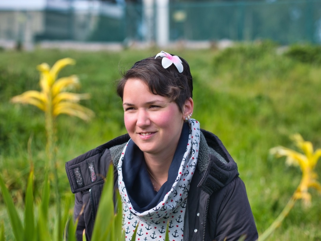 Karina, eine Frau mit kurzen, braunen Haaren und einer roten Blume im Haar sitzt auf einer Wiese. Im Vordergrund sind gelbe Blumen. Im Hintergrund grünes Gras.