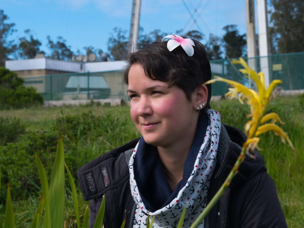 Karina, eine Frau mit kurzen, braunen Haaren und einer roten Blume im Haar sitzt auf einer Wiese. Im Vordergrund sind gelbe Blumen. Im Hintergrund grünes Gras.