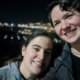 Caitlin, eine Frau mit langen, braunen Haaren, die zu einem Pferdeschwanz gebunden sind, lehnt ihren Kopf an Karinas Schulter. Karina ist eine Frau mit kurzen braunen Haaren. Beide Lächeln. Im Hintergrund sind die Lichter verschiedener Häuser zu sehen. Es ist Nachts.
