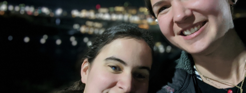 Caitlin, eine Frau mit langen, braunen Haaren, die zu einem Pferdeschwanz gebunden sind, lehnt ihren Kopf an Karinas Schulter. Karina ist eine Frau mit kurzen braunen Haaren. Beide Lächeln. Im Hintergrund sind die Lichter verschiedener Häuser zu sehen. Es ist Nachts.