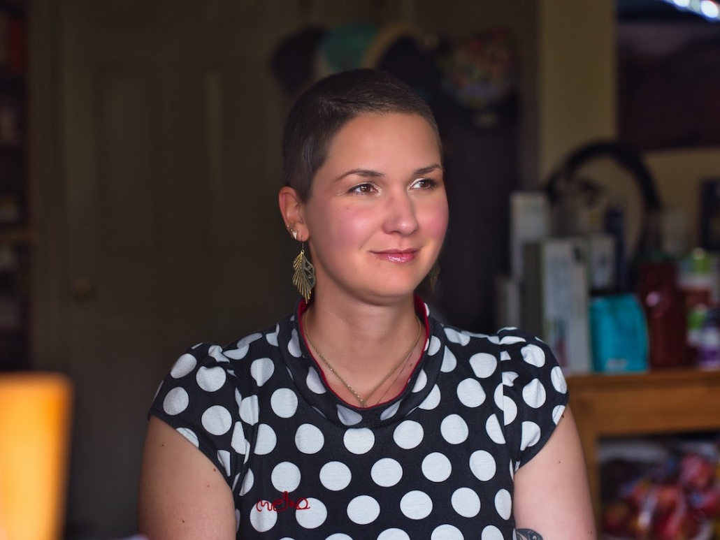 Karina, eine Frau mit 8mm rasierten haaren und langen federähnlichen Ohrringen sitzt auf einem Stuhl und schaut in die Ferne. Sie trägt ein Kleid mit Polkadots und lächelt.