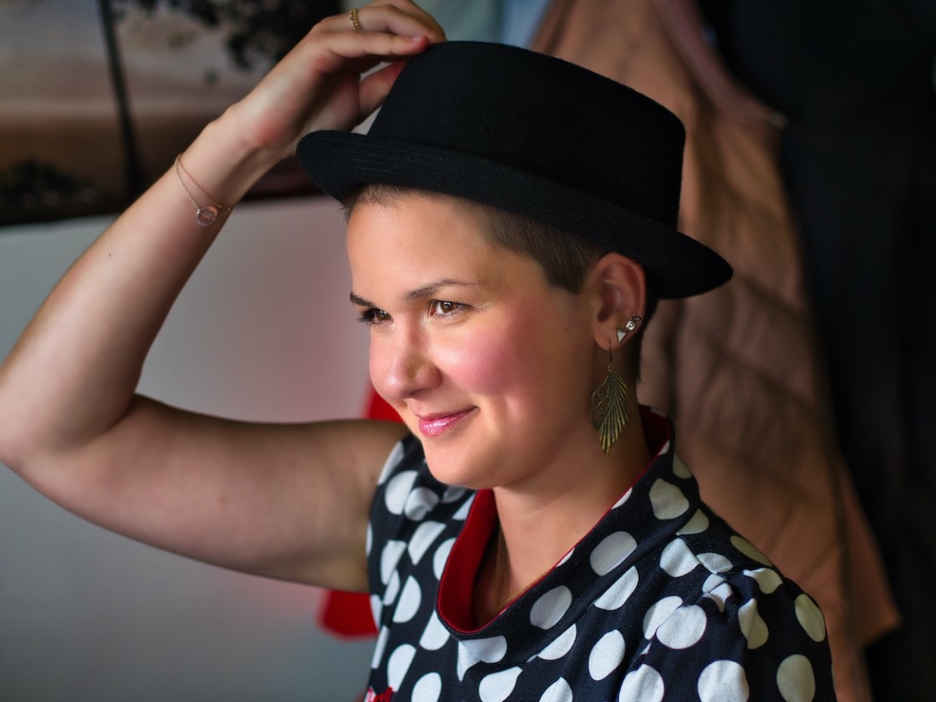 Karina, eine Frau mit 8mm rasierten Haaren und langen federähnlichen Ohrringen sitzt auf einem Stuhl und hebt mit ihrer rechten Hand einen runden Hut vom Kopf. Sie trägt ein Kleid mit Polkadots.