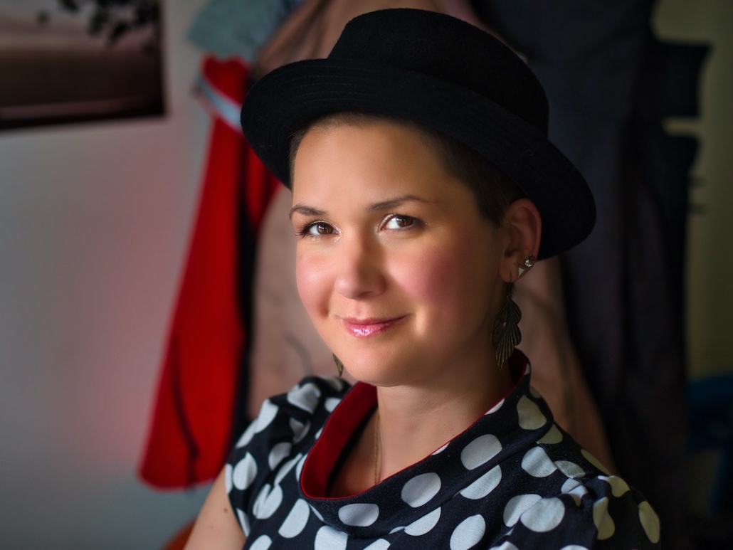 Karina, eine Frau mit 8mm rasierten Haaren und langen federähnlichen Ohrringen sitzt auf einem Stuhl und trägt einen runden Hut.