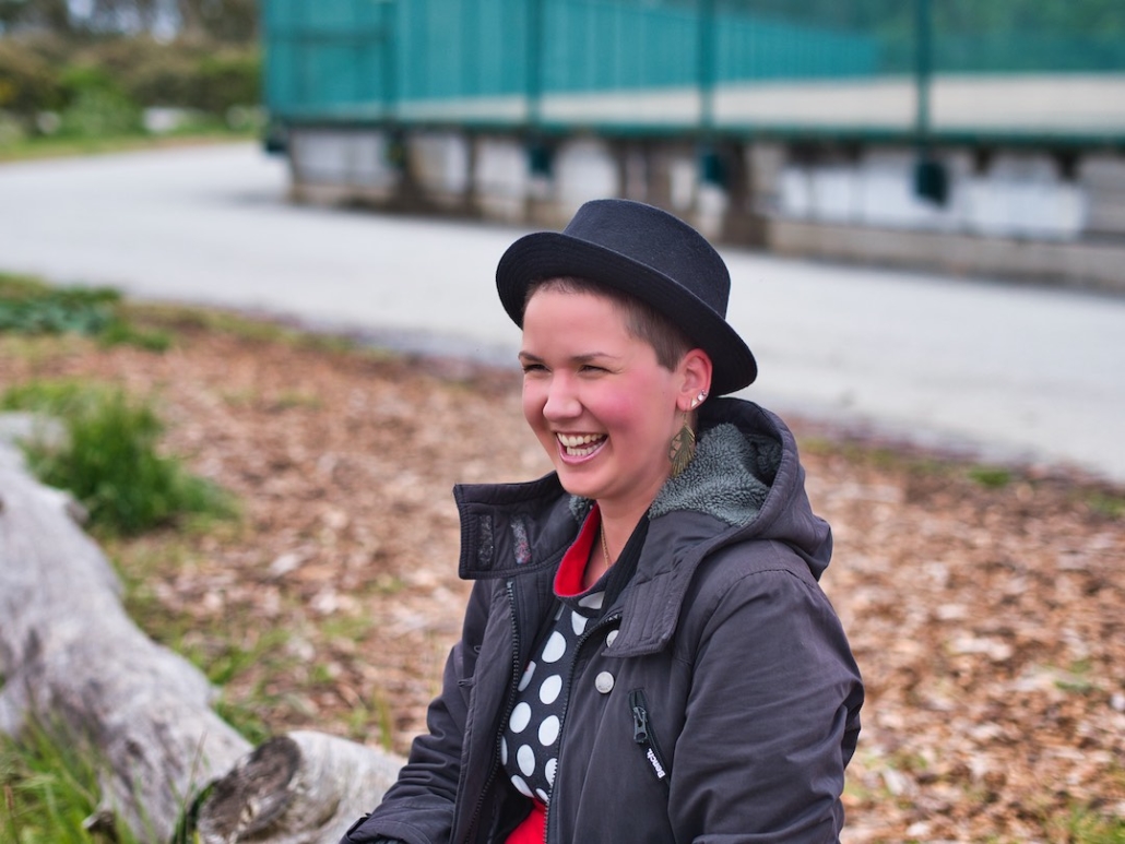 Karina, eine Frau mit 8mm rasierten Haaren und langen federähnlichen Ohrringen sitzt in der Natur auf einem langen Holzbalken und trägt einen runden Hut. Sie lacht herzhaft.