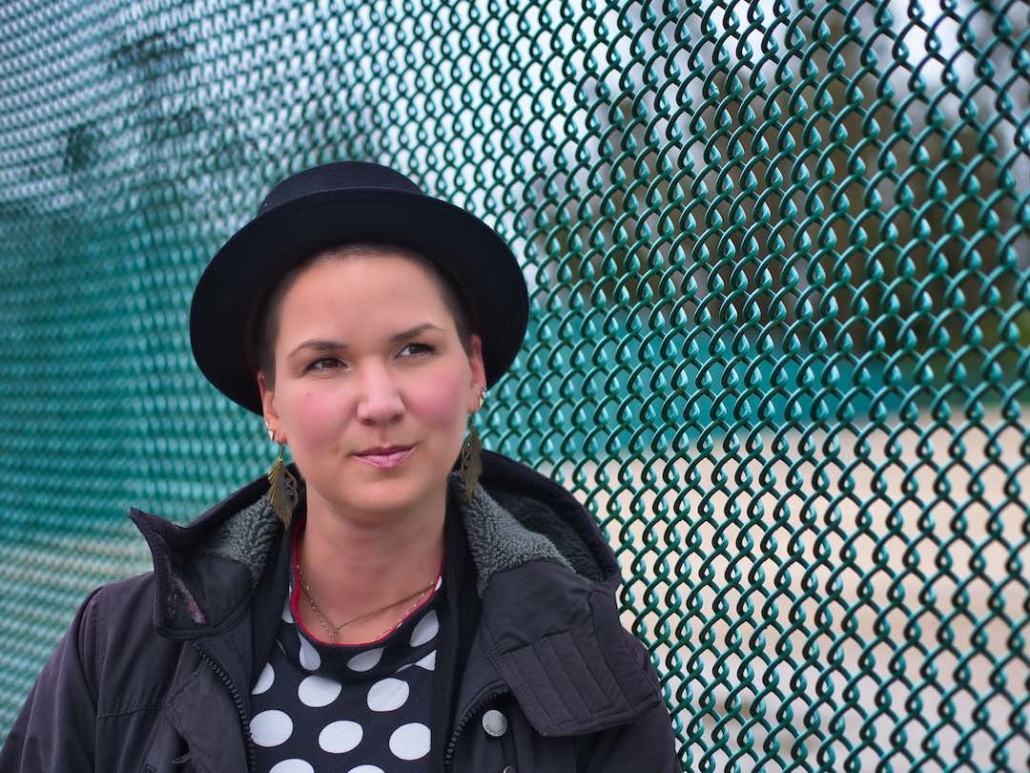 Karina, eine Frau mit 8mm rasierten Haaren und langen federähnlichen Ohrringen trägt einen runden Hut. Sie steht an einem grünen Zaun.