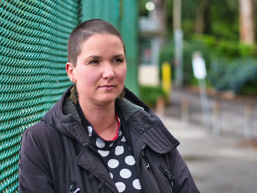 Karina, eine Frau mit 8mm rasierten Haaren und langen federähnlichen Ohrringen steht an einen grünen Zaun angelehnt.