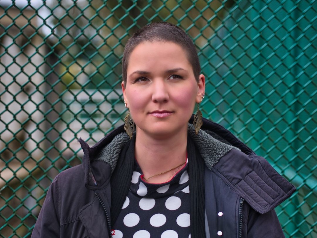Karina, eine Frau mit 8mm rasierten Haaren und langen federähnlichen Ohrringen steht an einen grünen Zaun angelehnt.