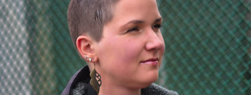Karina, eine Frau mit 8mm rasierten Haaren und langen federähnlichen Ohrringen steht an einen grünen Zaun angelehnt.