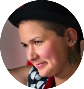 Karina, eine Frau mit rasierten Haaren und schwarzem Hut in der Seitenansicht. Sie trägt große Federartige Ohrringe und Lippenstift.