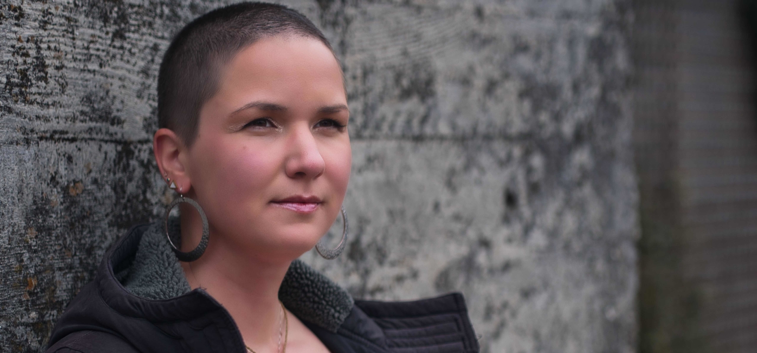 Karina, eine Frau mit 7mm langen Haaren lehnt an einer Wand. Sie trägt große runde Ohrringe und Lippenstift.