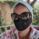 Karina, eine Frau mit kurzen braunen Haaren trägt ein blaues Stirnband, eine schwarze Maske und eine Sonnenbrille.