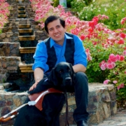 Belo, a young man with short brown hair sits on beautiful stairs in a garden covered in red roses. He smiles and has one hand on his black guide dog