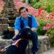 Belo, a young man with short brown hair sits on beautiful stairs in a garden covered in red roses. He smiles and has one hand on his black guide dog