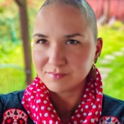 Karina, eine Frau mit fast rasiertem Kopf steht in ihrem Garten. Sie trägt ein Shirt mit Polka Dots und einen Sweater mit Totenköpfen