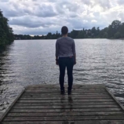 A woman stands on a wooden walkway of a lake and looks into the distance with a threatening dark and cloudy sky above