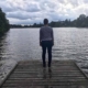 A woman stands on a wooden walkway of a lake and looks into the distance with a threatening dark and cloudy sky above