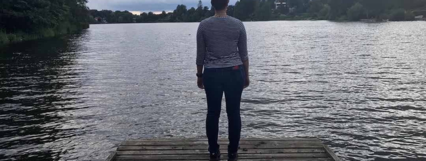 A woman stands on a wooden walkway of a lake and looks into the distance with a threatening dark and cloudy sky above