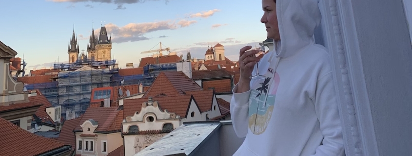 Karina, a woman with short brown hair leans against a wall on a balcony during sunset. She wears a white hoodie and holds a glass of wine in her hand. In the background you can see an old church and many older houses.